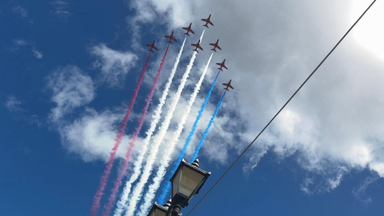 The photo shows 8 red arrows planes mid-flight in an arrow-shaped formation. Two are releasing red smoke behind them, three are releasing white smoke, and to are releasing blue smoke. 