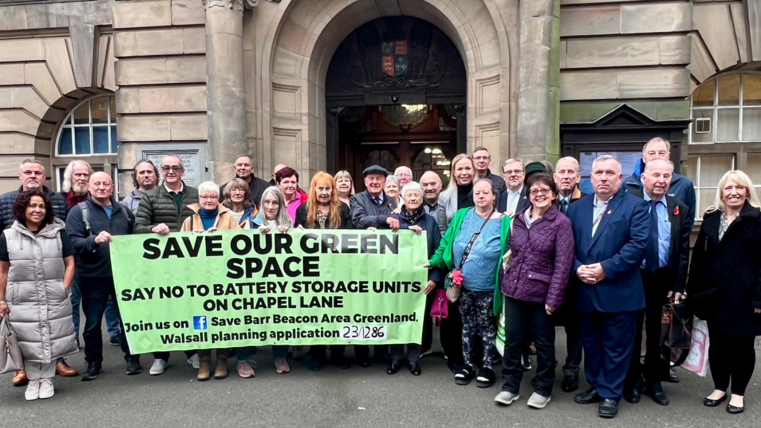 Dozens of men and woman stand outside the council building with some holding a banner that says "Save our green space - say no to battery storage units"