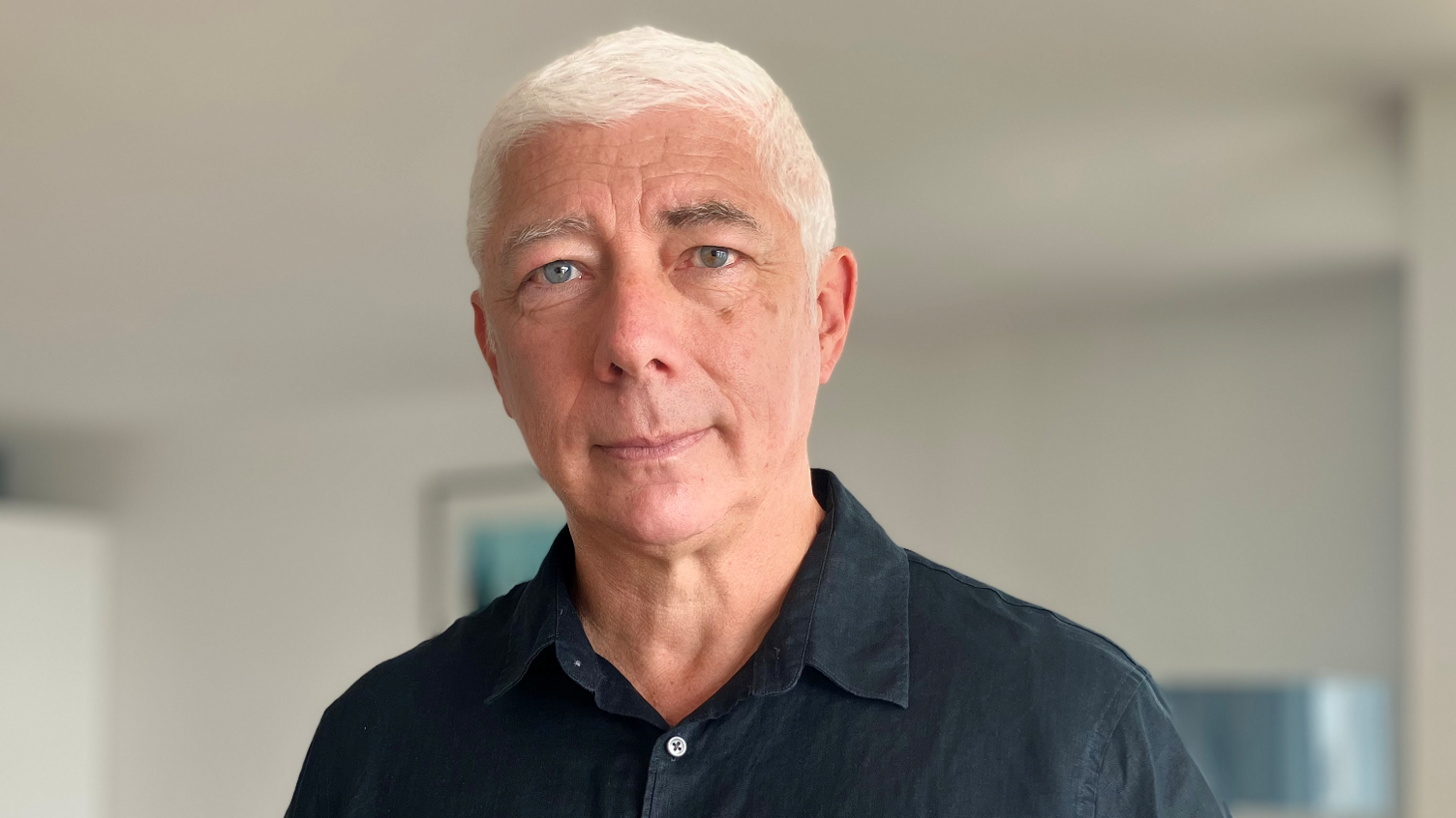 David Rogers is wearing a black buttoned shirt, with the top button undone, and has white, short hair.
