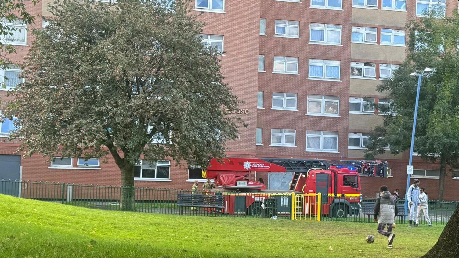 A red fire service vehicle with what appears to be a long-reach ladder on it parked outside the building. The ladder is down and in the foreground a couple of children wearing coats can be seen kicking a football. 