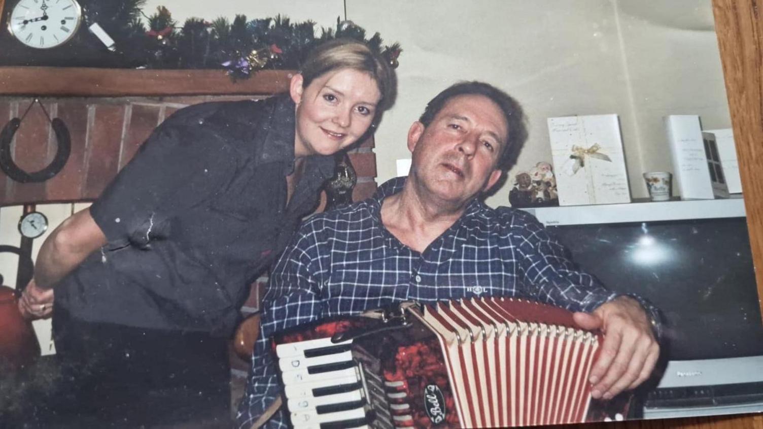 An old family photo of Lisa Allen with her father Ken Hopkins. He is wearing a navy and white checked shirt aqnd is holding a red and black accordion. She is leaning close to her father and smiling at the camera. She has blonde hair tied back from her face and is wearing a dark shirt.