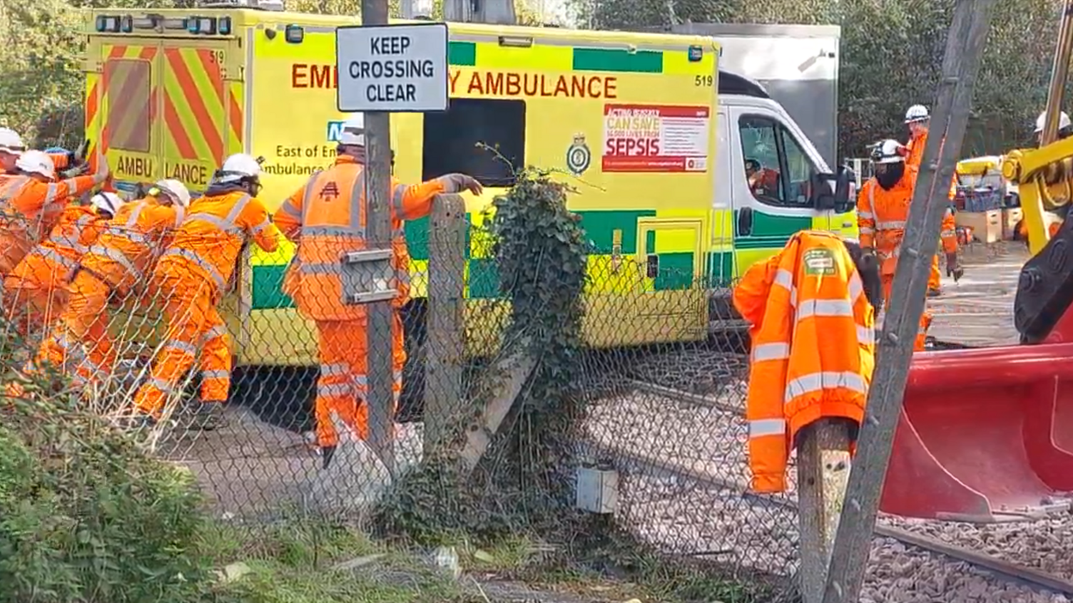 A group of maintenance workers wearing orange hi-vis clothing pushing an ambulance from behind over a railway crossing.