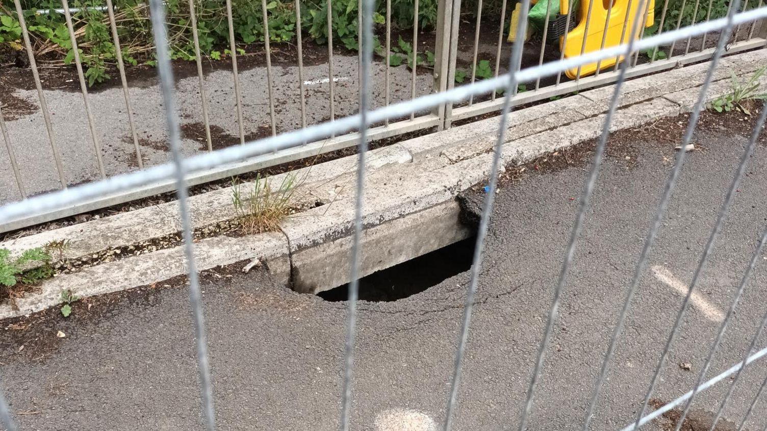 A sinkhole can be seen on the side of a pavement through a metal fence. It appears to be somewhere between 0.3m (1ft) and 0.6m (2ft) long and about 15cm (6ins) and 0.3m (1ft) wide.