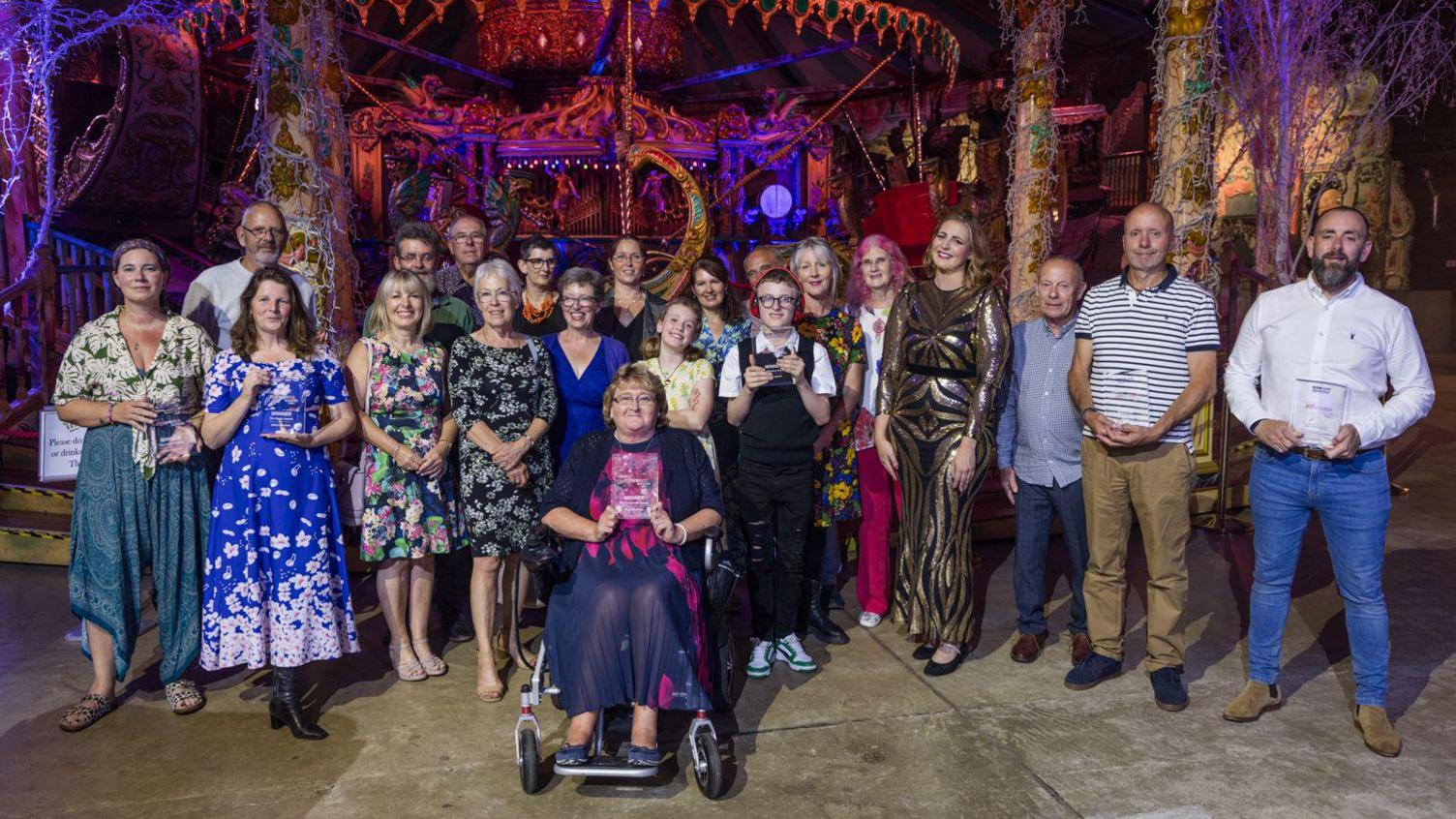 A group of people standing and one person sitting in a wheelchair. A number are holding their awards made of glass while smiling for the camera.