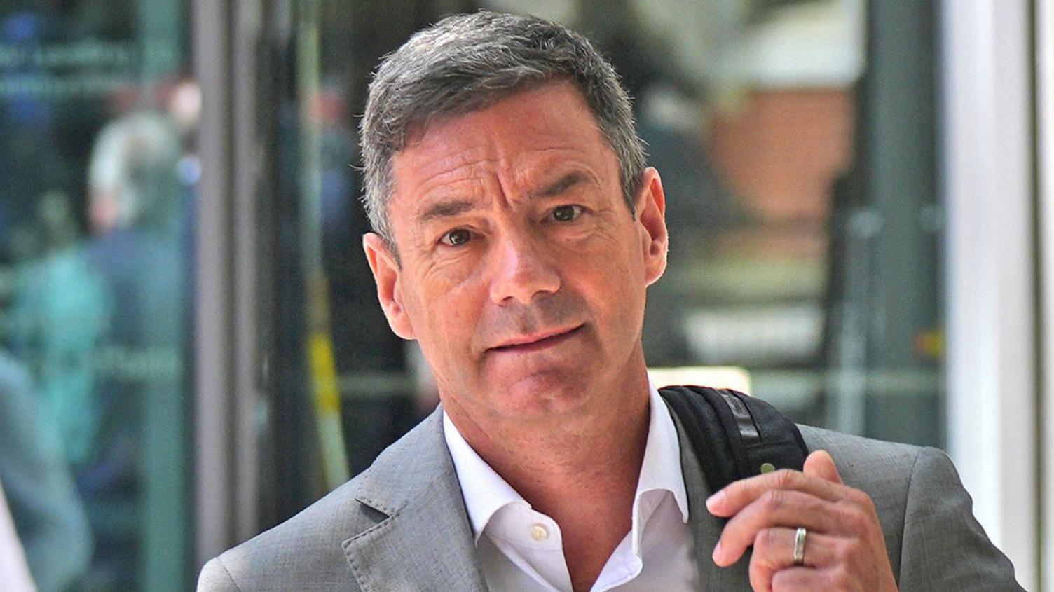 Lawrence Gosden has short grey hair and is wearing a grey blazer and white shirt, whilst carrying a backpack.