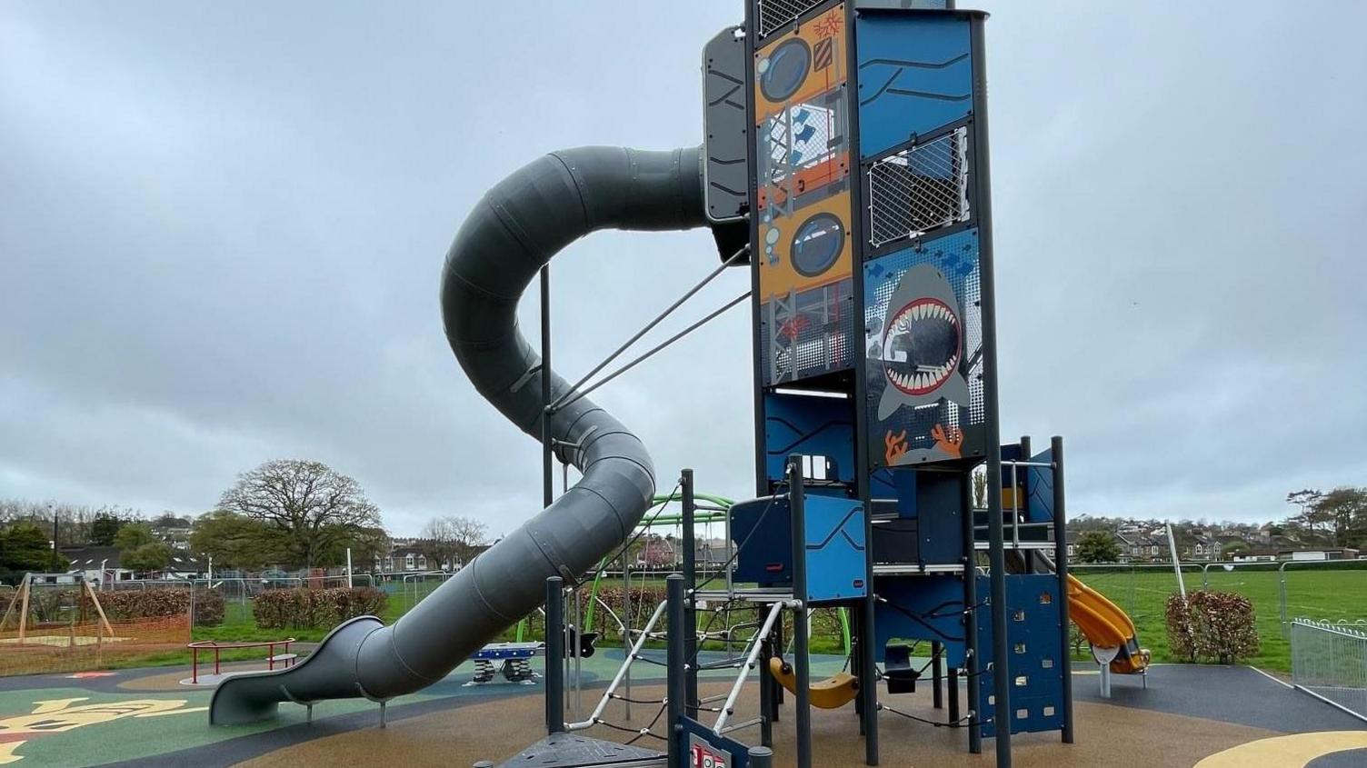 Tall play tower with different activities. There is a grey slide with a twist and grass in the background.