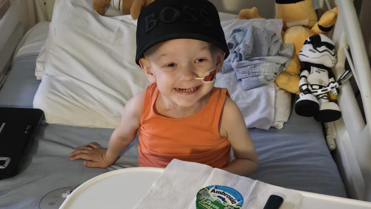 The photo shows Jonah sitting in a hospital bed. He has a nasogastric tube inserted and he is wearing an orange vest and black hat. He is smiling. In front of him on a hospital table is a pot of food which is unopened.