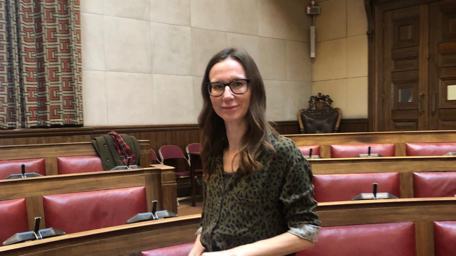 Harriet Morgan in the council chambers. She has long brown hair and is smiling at the camera. She is wearing a green leopard print shirt. 