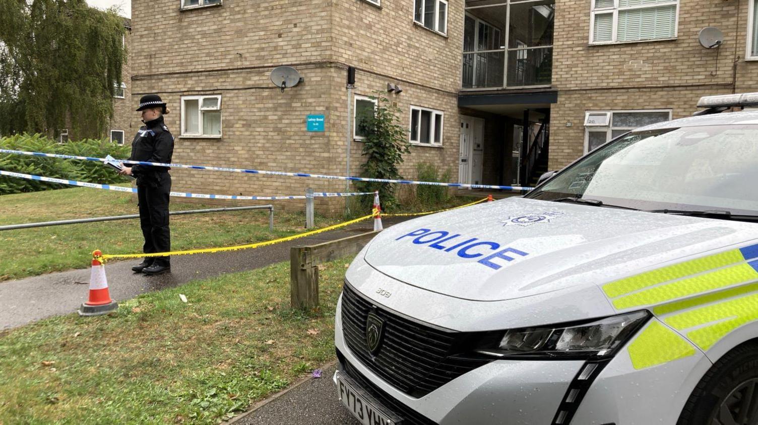 A police officer standing outside a cordoned off three-storey block of flats with a police car in the foreground.