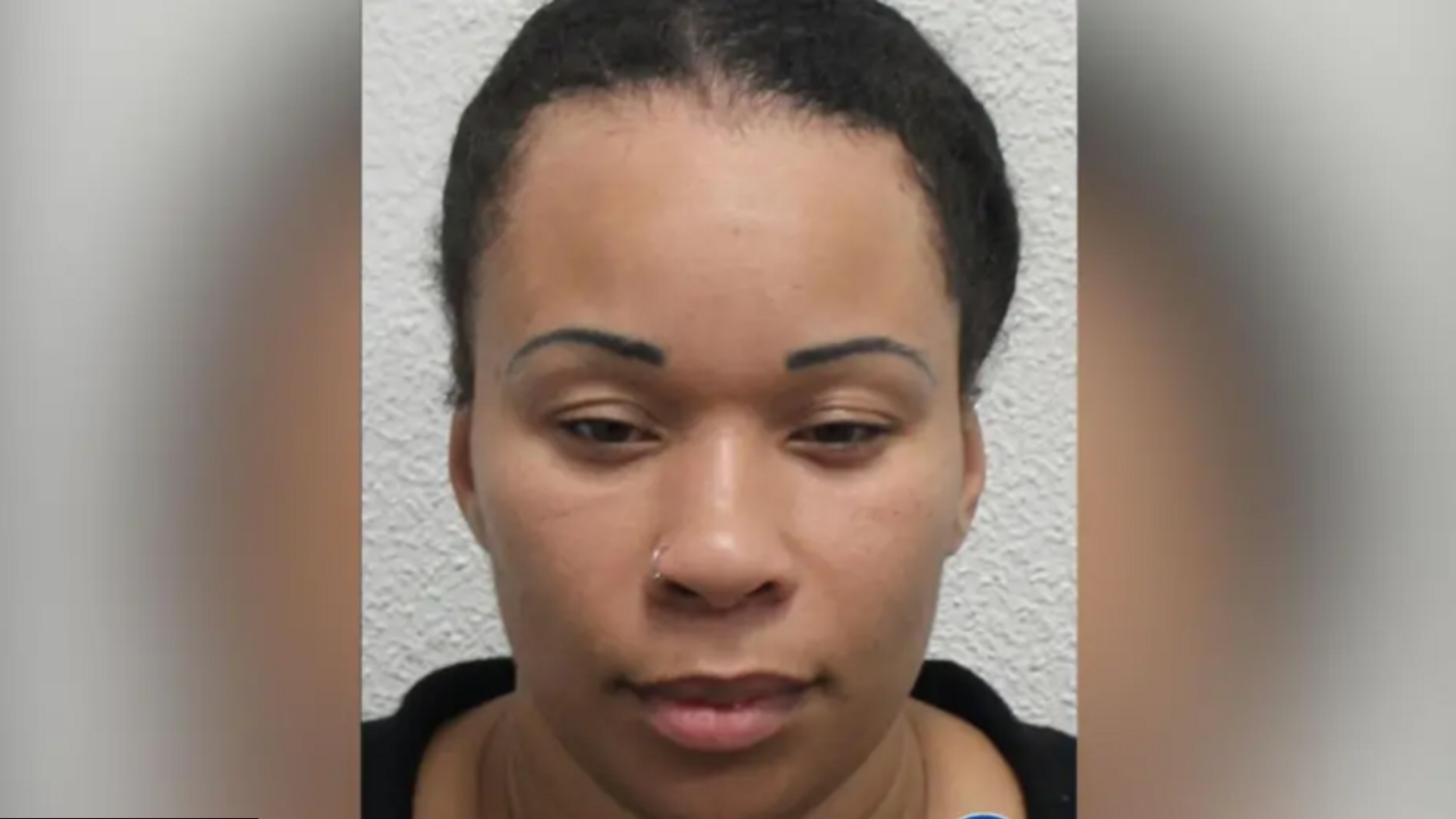 A woman with pulled-back black curly hair looking at the camera in a police mugshot. She is wearing a black top and small gold nose ring.