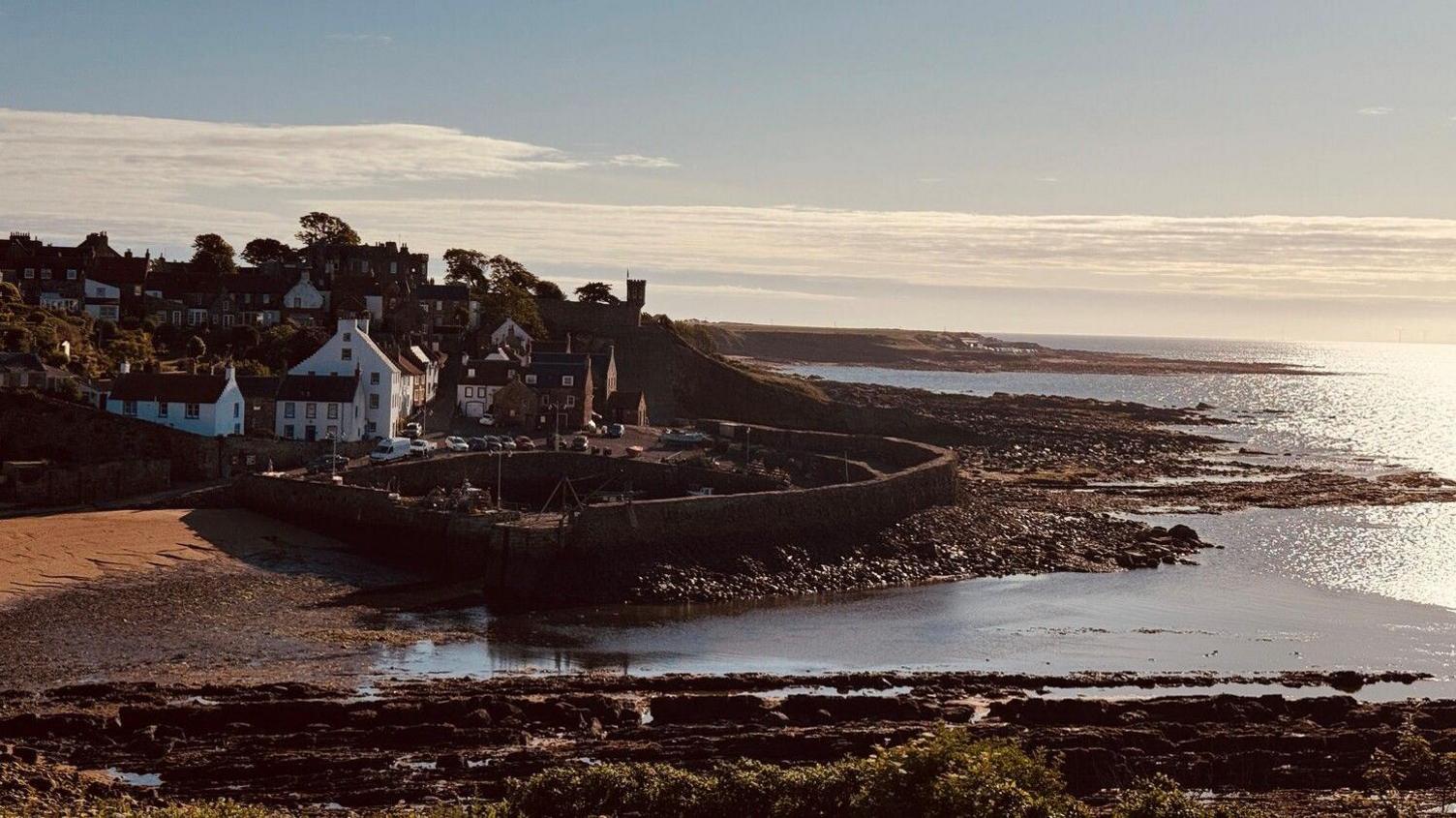 A coastal town. The sun shines on the water with a small sandy beach and rocks by the coast. The little town can be seen in the distance.