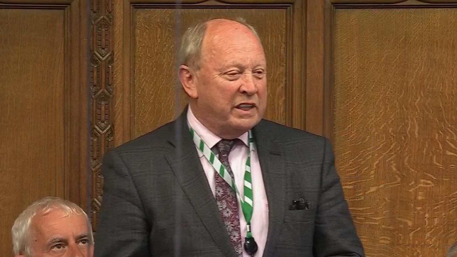 Jim Allister speaking in the House of Commons wearing a suit and tie and green lanyard