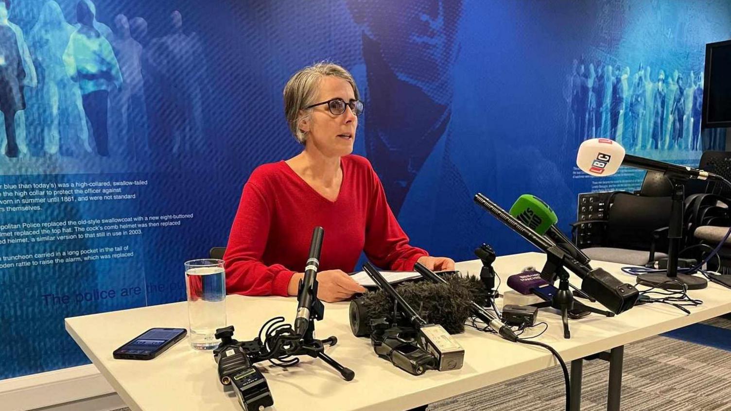 Detective Chief Inspector Charlotte Whalley holds a statement while sat on a table filled with microphones, with a glass of water nearby. The DCI is wearing a red top and black glasses. 