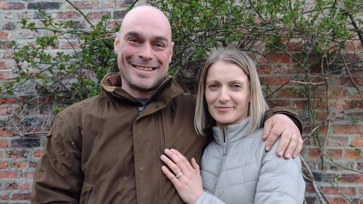 Dorota Spilman pictured with her husband. He is bald with blue eyes and is wearing a brown jacket, while Dorota has shoulder-length blonde hair. Both of them are smiling in the photo.