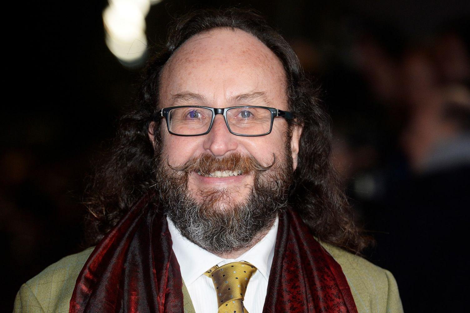 Dave Myers, photographed arriving at the National Television Awards 2015. He is balding but has long hair at the back, square glasses, a greying beard and a waxed moustache. He is wearing a greenish jacket, a white shirt and a spotted tie to match the jacket. He also is wearing a dark red silk scarf. 