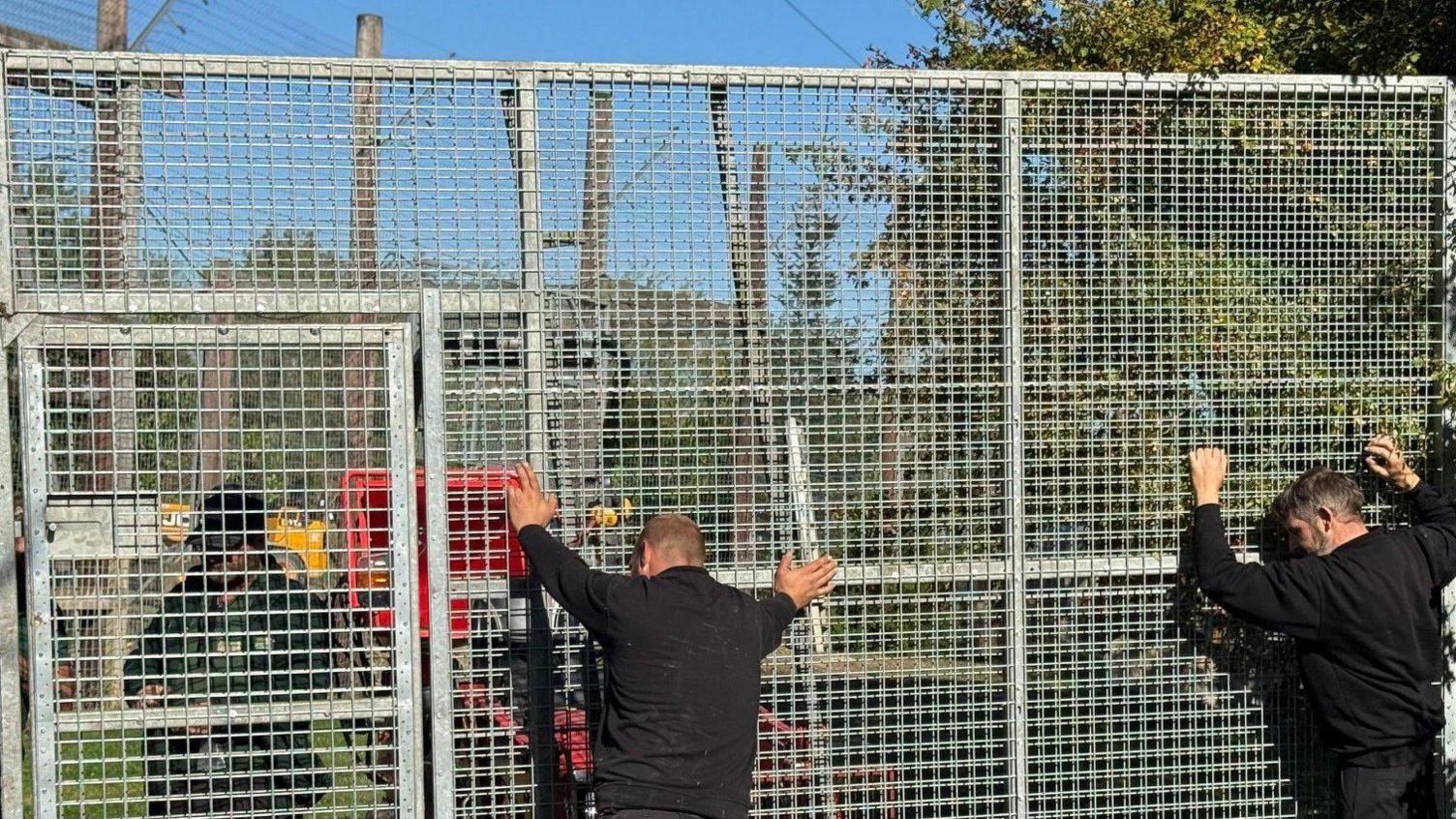 A tall metal gate with square holes in it with two men dressed all in back leaning their hands against it.