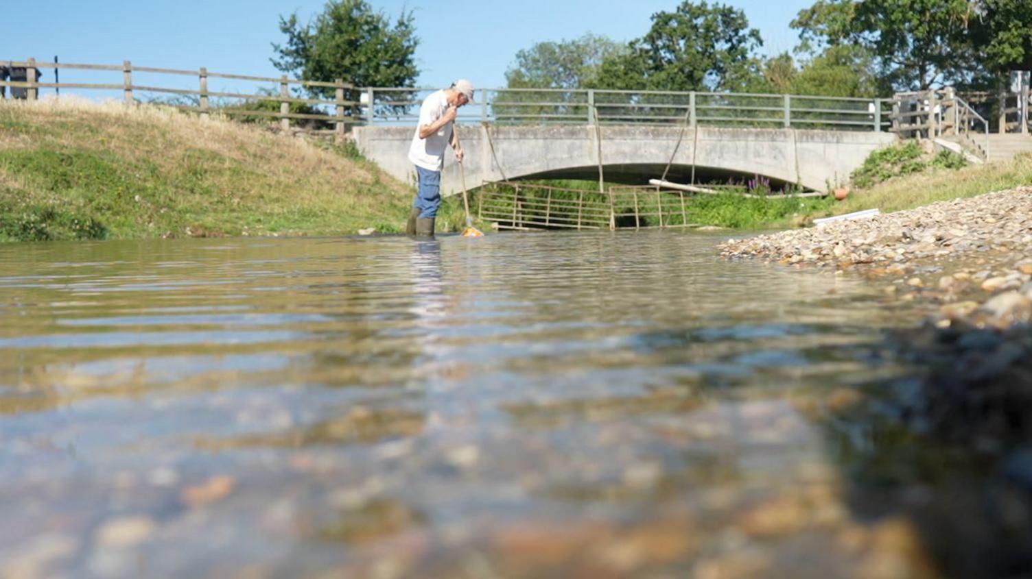 A man stands in a river with the water up to his thighs. He is wearing wellington boots. The water stretches out from the camera and a bridge is in the background.