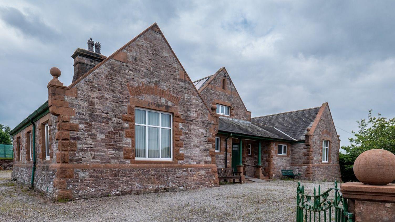 The front of the Toppin Memorial Hall, the village hall at Skelton, near Penrith, Cumbria. It is a large double-fronted village hall made of local red sandstone, with an old stone wall and painted green metal gates in front of it, a gravel car park and has a porch way entrance. The new solar panels are visible on the roof, and the sky above is grey. 