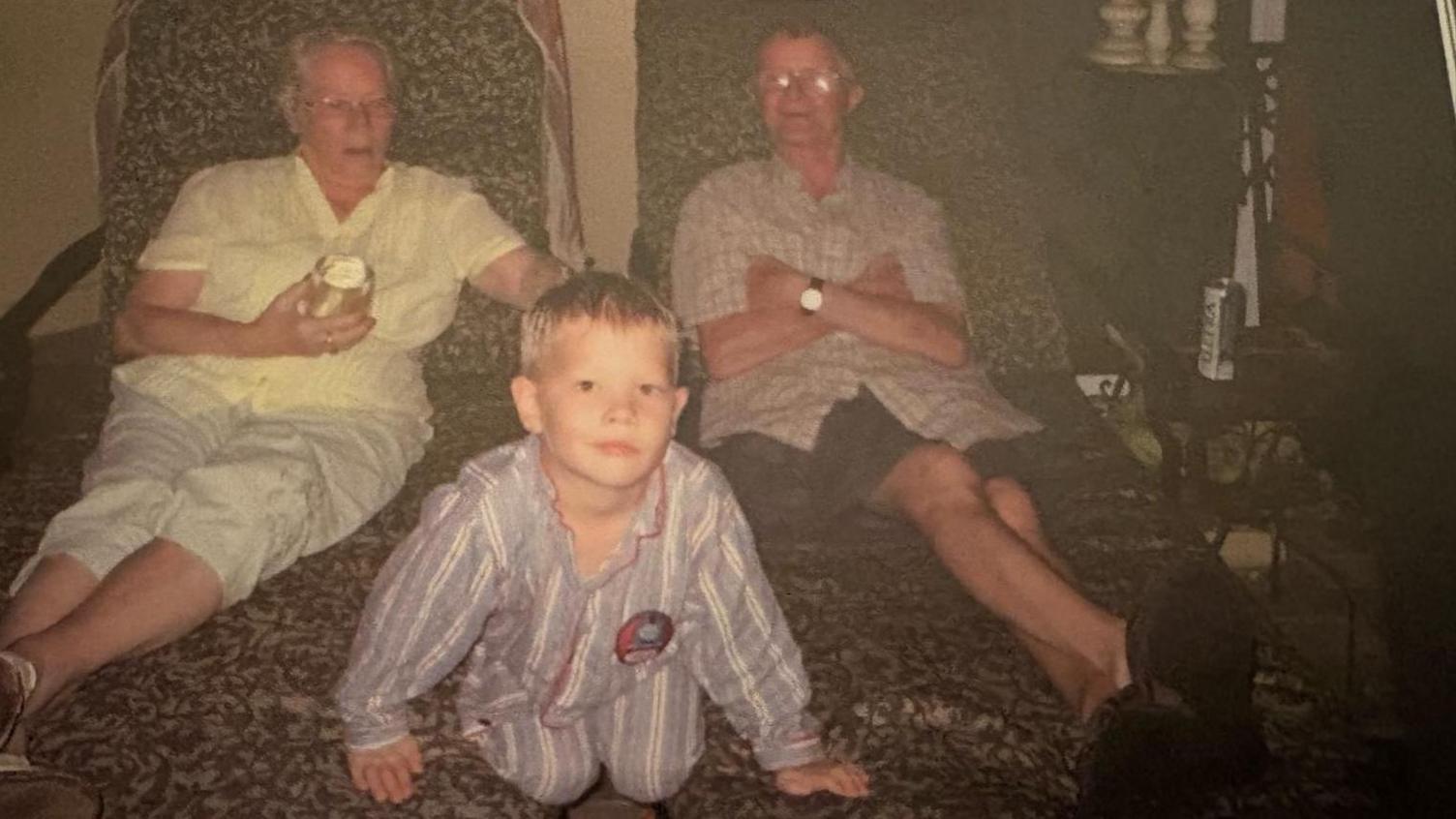  A faded family photograph in which Charlie as a young child is wearing Thomas the Tank Engine pyjamas. Charlie's grandparents are sitting on sun loungers behind him.