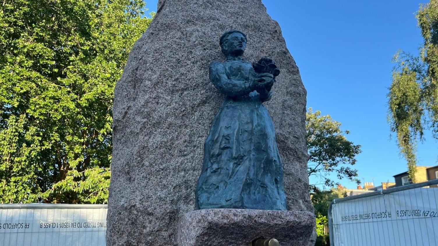 A statue of a woman on a large rock-style water fountain. There are tall trees in the background and the sky is clear blue.