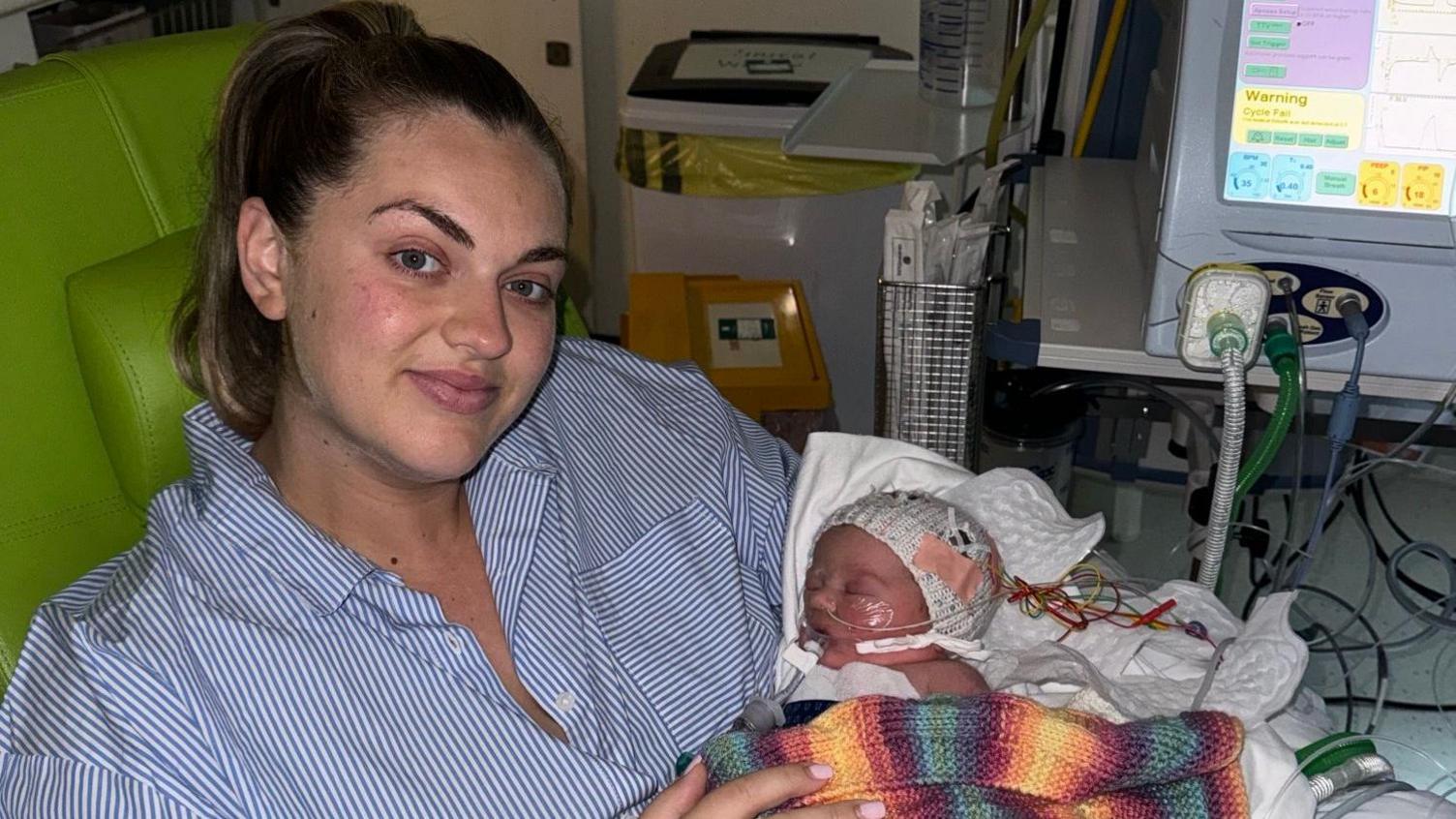 A woman with blonde hair, wearing a blue and white striped shirt, holds a newborn baby wrapped in a multicoloured blanket. There are tubes connecting the baby girl to a machine.