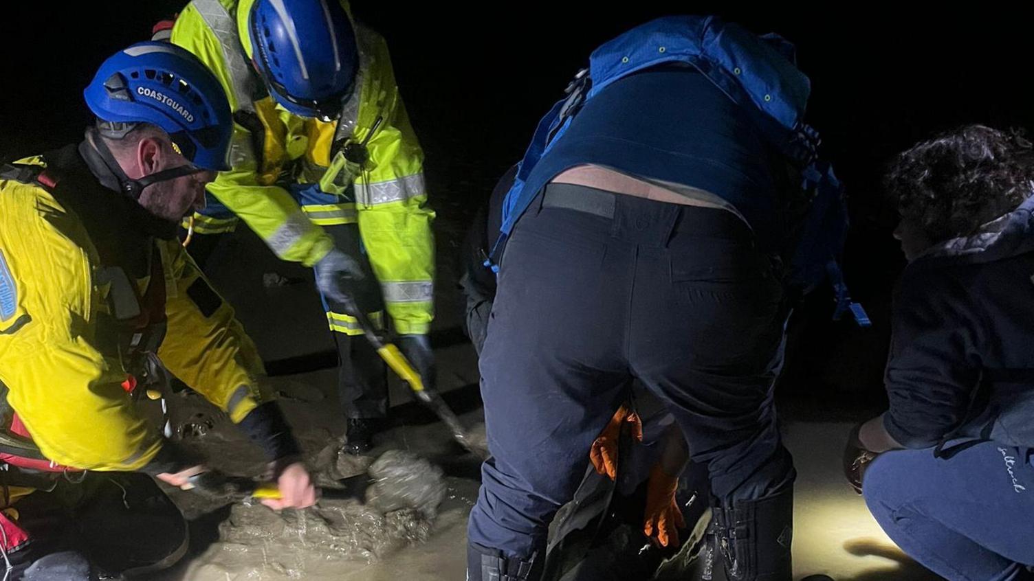 Three men are digging in the sand with a fourth man crouched down, It is dark but there are torchlights illuminating where they are working 