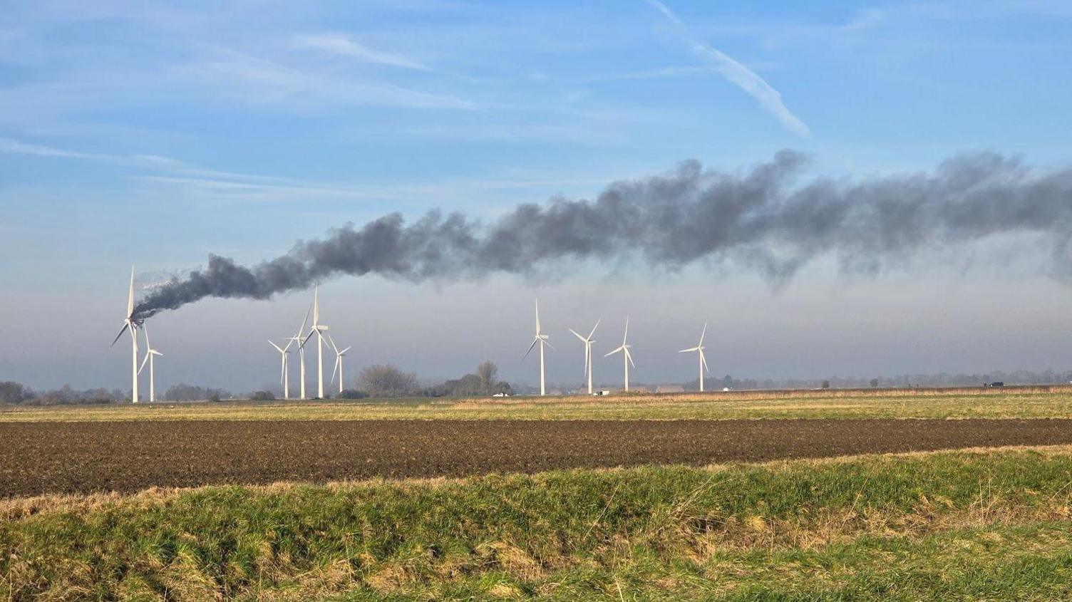 Smoke is billowing from a wind turbine that is on fire in a field. There are nine turbines nearby. The sky is blue and there is a field in the foreground that is muddy