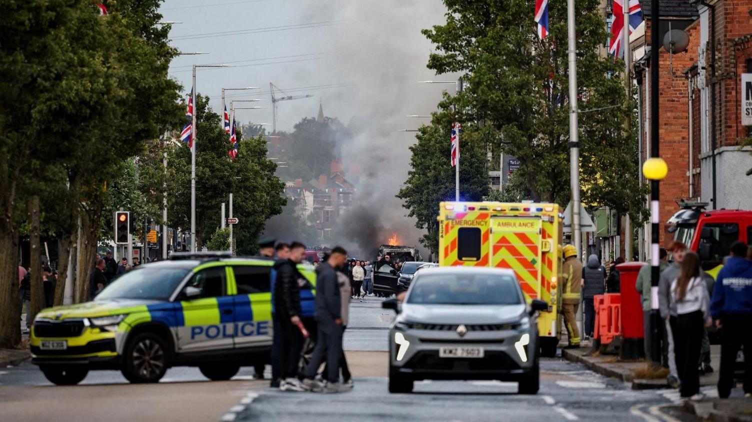 Police vans and ambulance parked with people walking in the foreground and disorder in the background at a distance