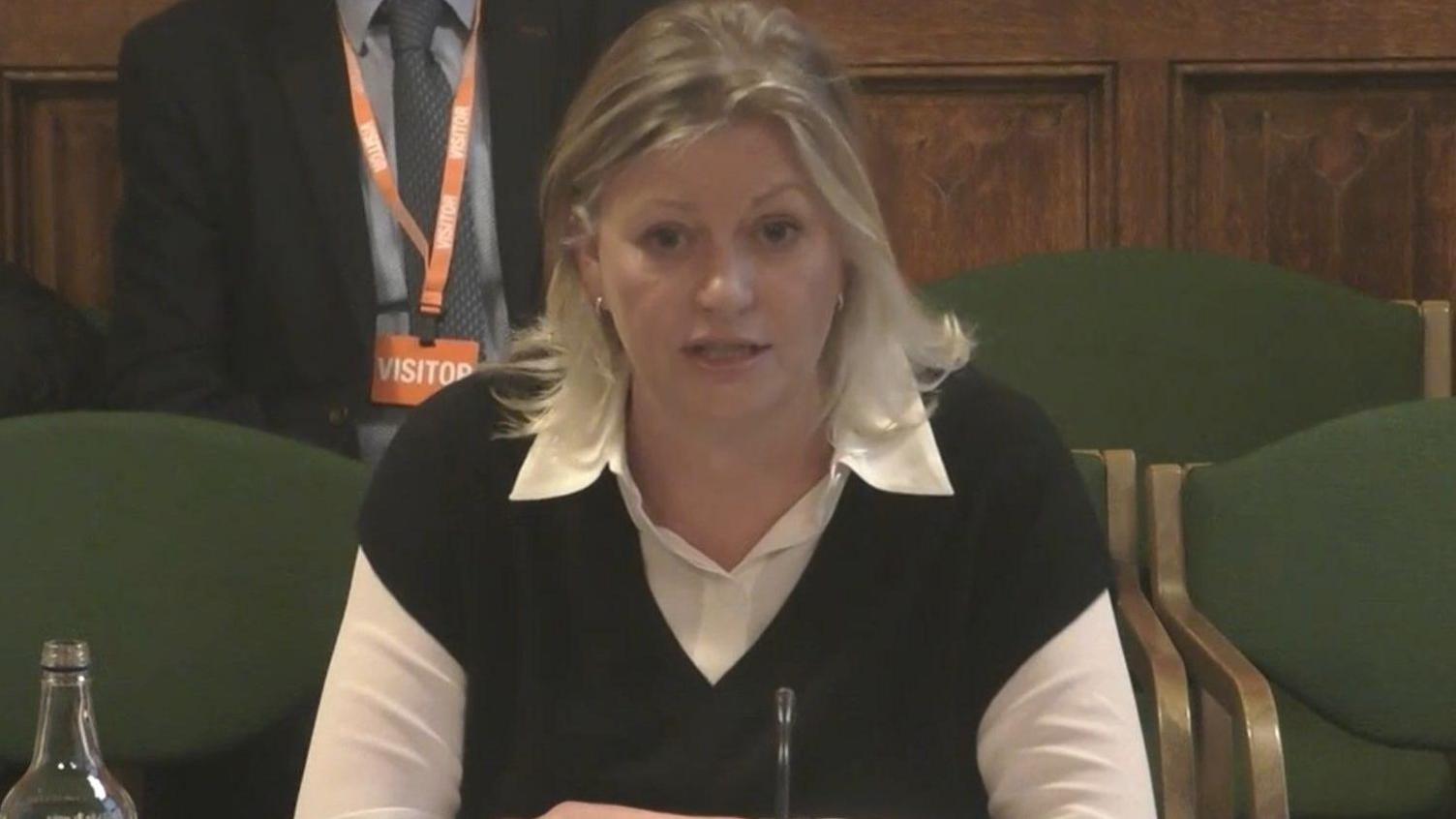 A woman with shoulder length blonde hair mid speech wearing a white shirt with a black vest over the top.