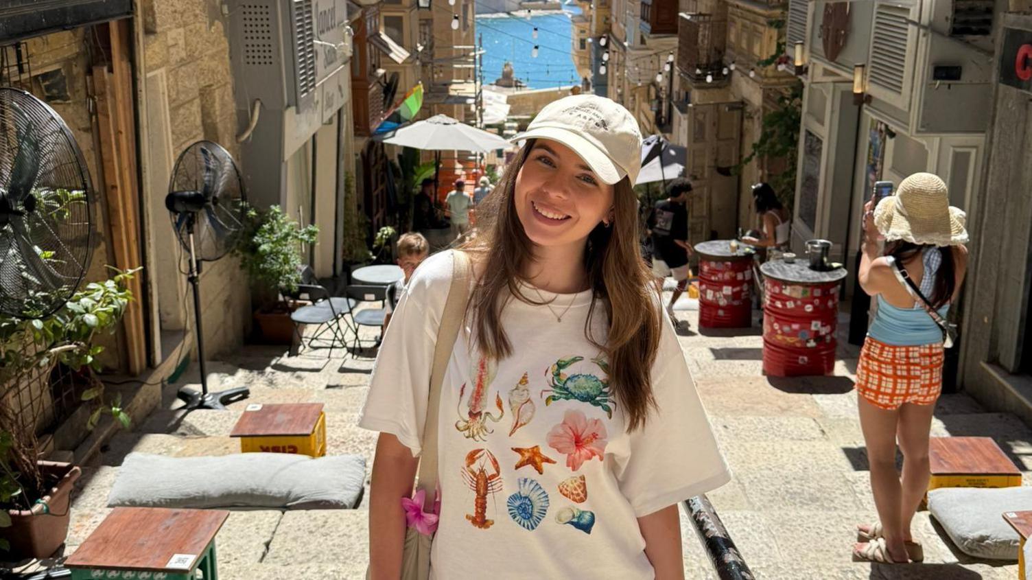 A young woman stands in a narrow, sunny street that slopes down towards a harbour area in the background. There are seats and tables outside most of the buildings and black electric fans can also be seen. To the right, a woman with her back to the camera takes a photo on her phone.