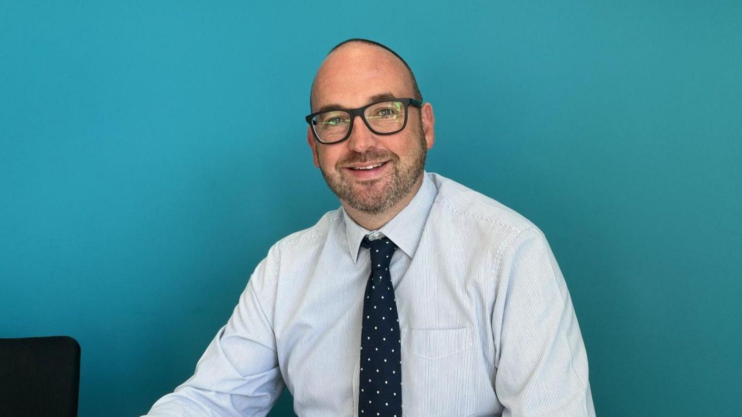 A man smiling into camera wearing a shirt and a polkadot tie 