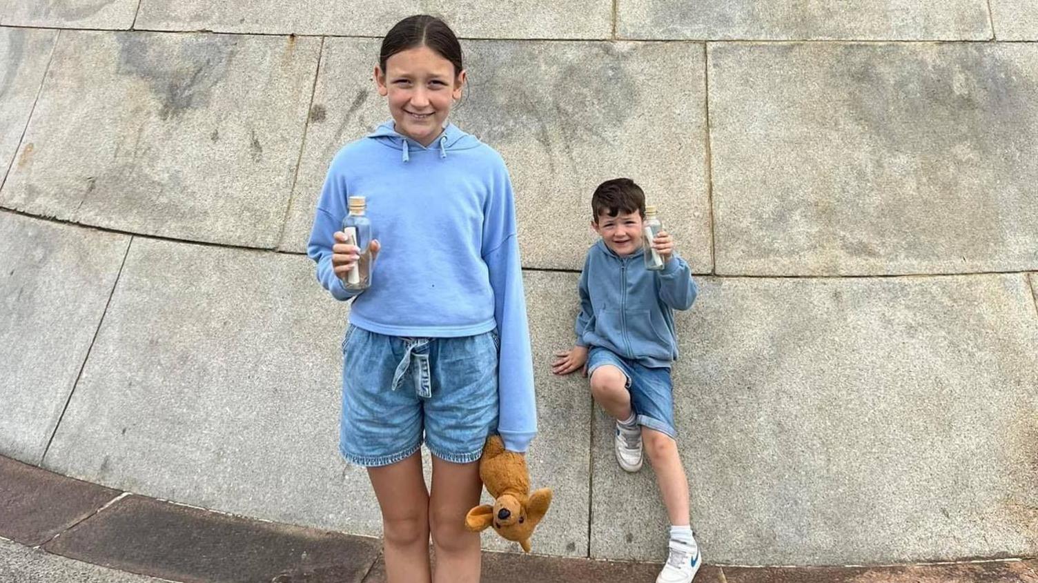 Grace and Harry holding their messages in a bottle and smiling. They are both wearing blue hoodies and shorts. Grace is holding a stuffed toy.