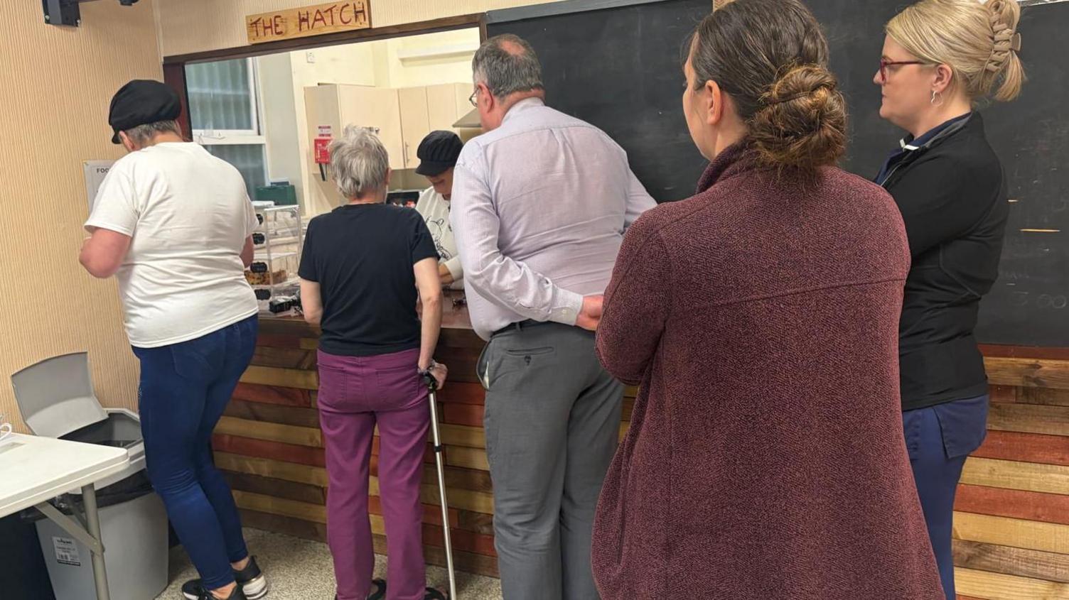 Five people stand in line at a kitchen hatch with the sign "The Hatch" above it.