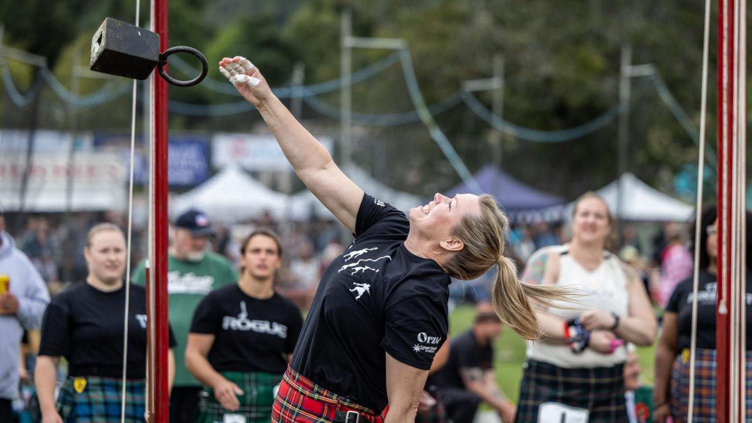 An athlete leans back as she throws a weight, trying to get it as high as she can. The athlete is wearing a black t-shirt and a red tartan kilt. She is watched by other competitors.