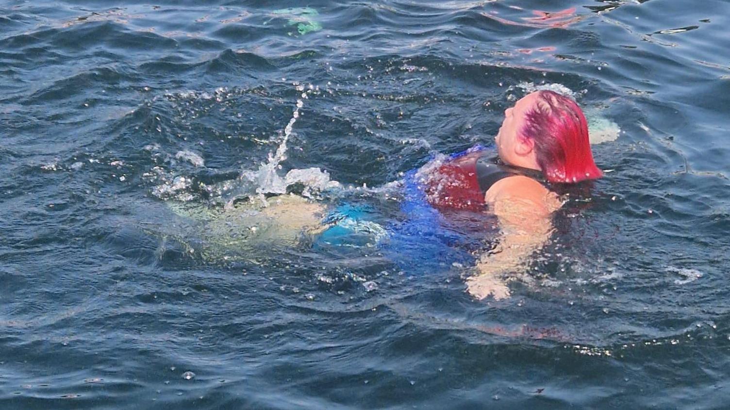 A woman with pink hair is floating on her back in water with her arms outstretched.