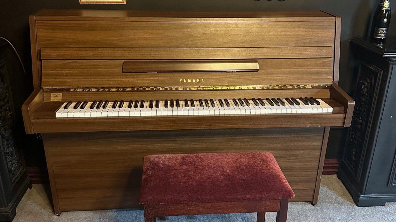 A brown wooden Yamaha upright piano with the lid open. There is a red velvet covered piano stool next to it and it sits in a dark room with a silvery coloured carpet.