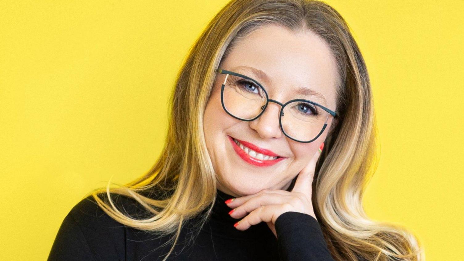 A photo of a blonde woman in a black jumper stood in front of a bright yellow background. She is wearing glasses and smiling at the camera