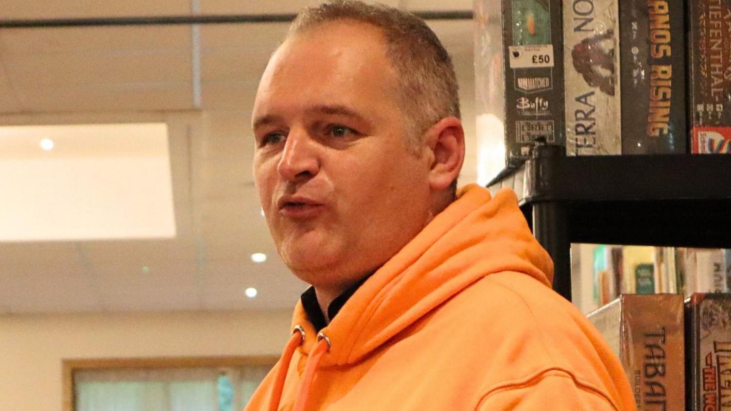 Paul Harris is stood near a shelf full of board games and wearing an orange hoody. He has short grey hair.