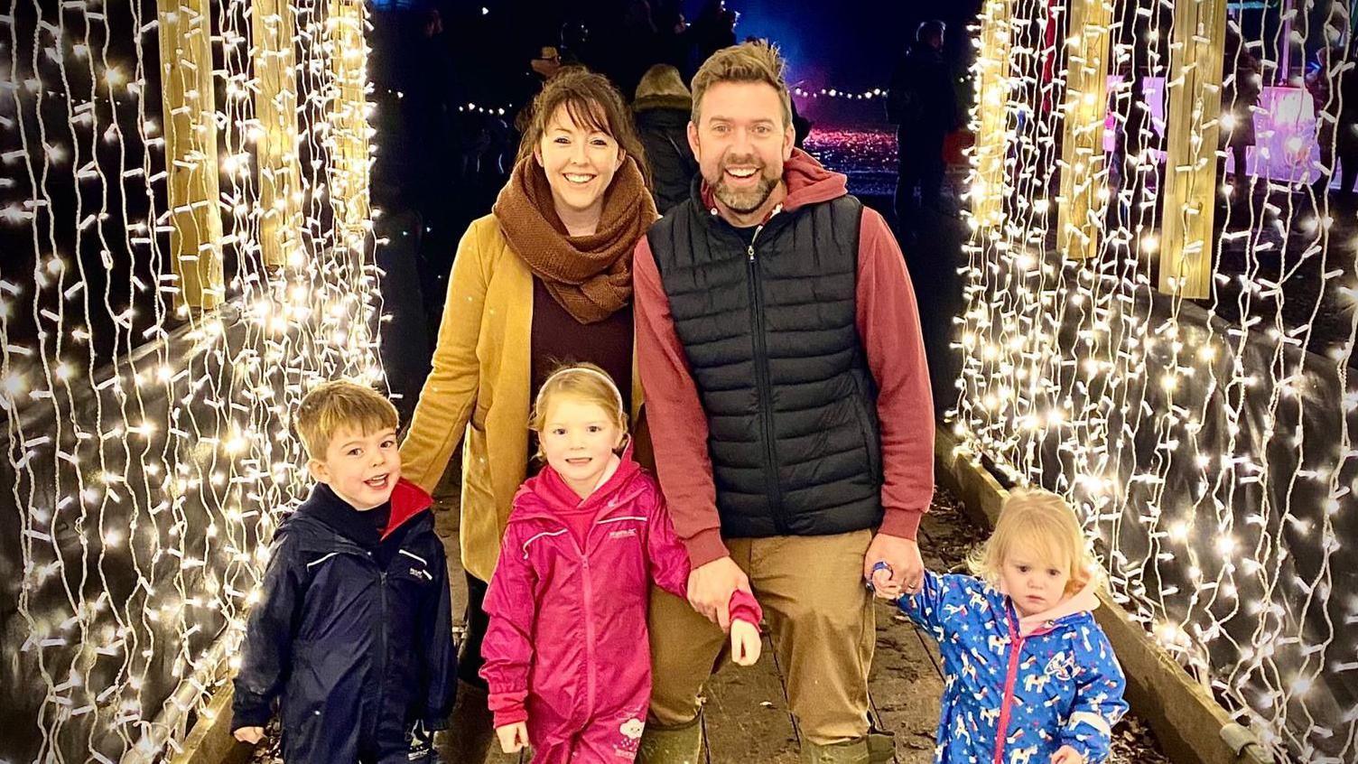 The family stands under an archway of fairy lights. Katie and Tom are with their three children, a boy and two girls.