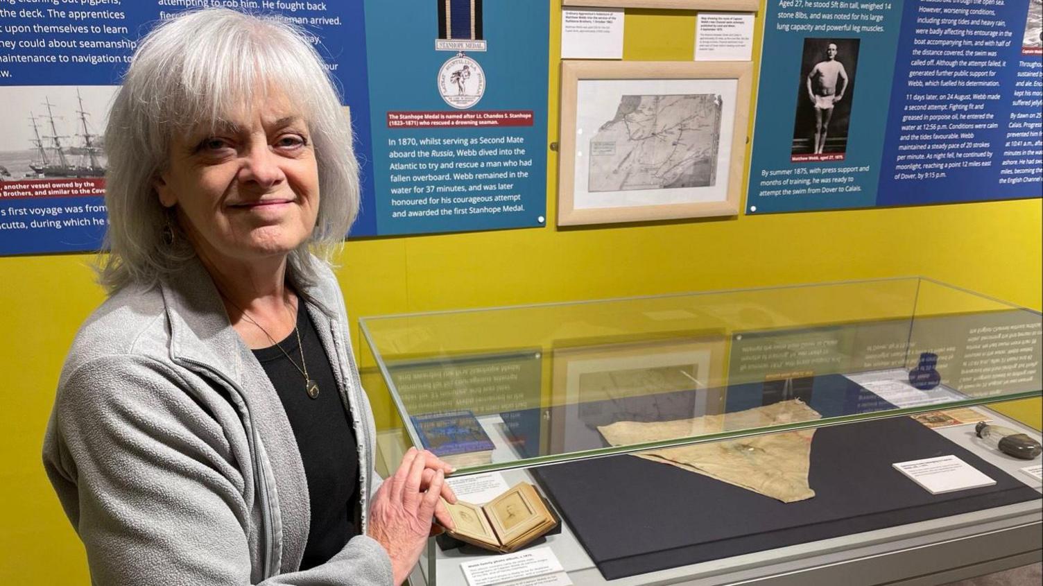 A woman with shoulder-length grey hair wears a grey jacket and black top with a gold necklace. She is smiling at the camera and standing next to a display case featuring a pair of beige briefs on a black background. Behind her is a blue display board bearing information about Caption Matthew Webb.