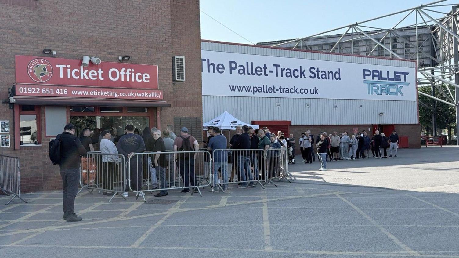 The word ticket office is above windows on the outside of the stadium on the left of the photo. The queue starts there, with people behind barriers, and continues towards the right of the image outside by the club's main stand.