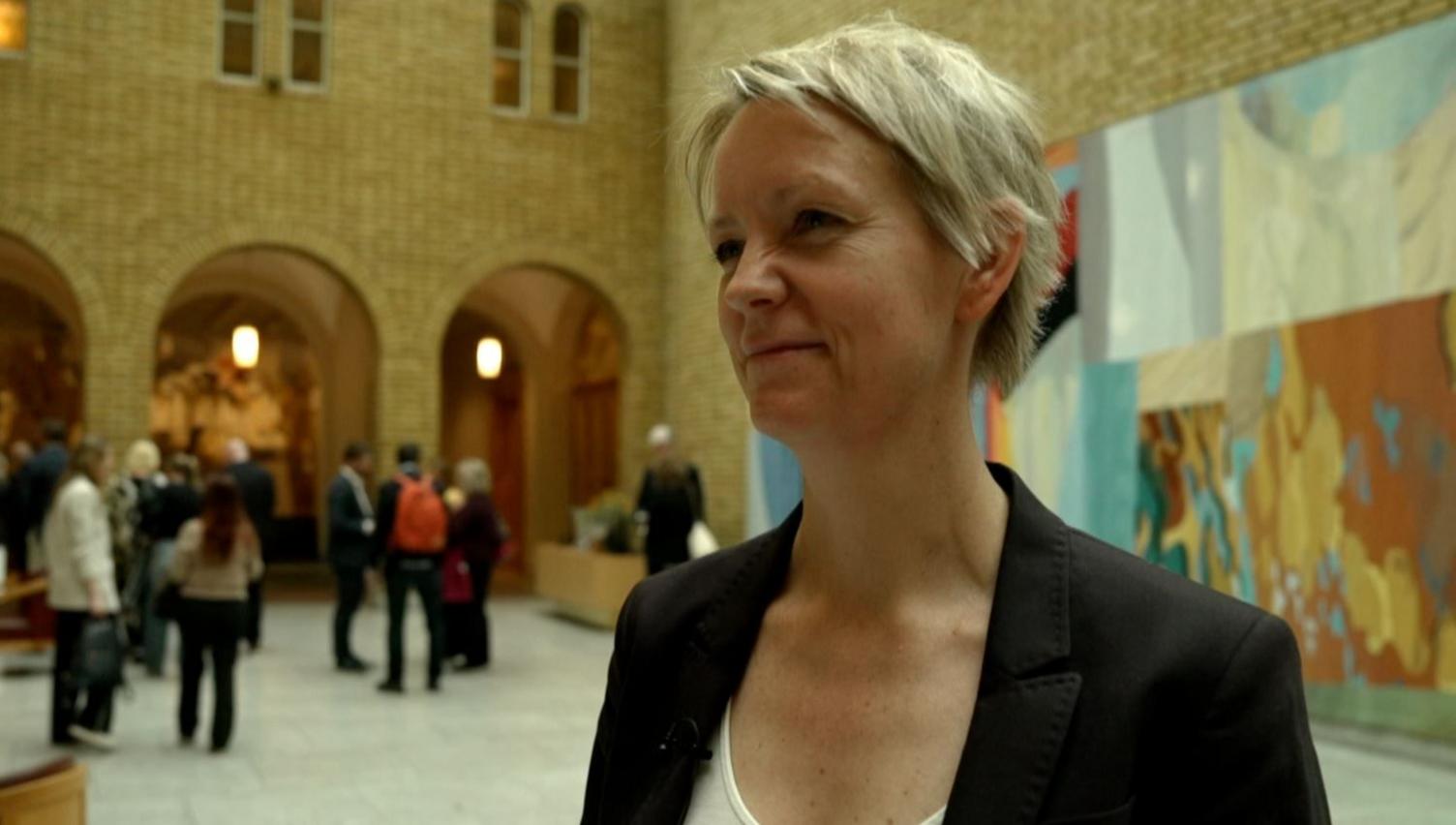 Ingrid Fiskaa in a large hall. She has short blonde hair and is wearing a white shirt and black jacket