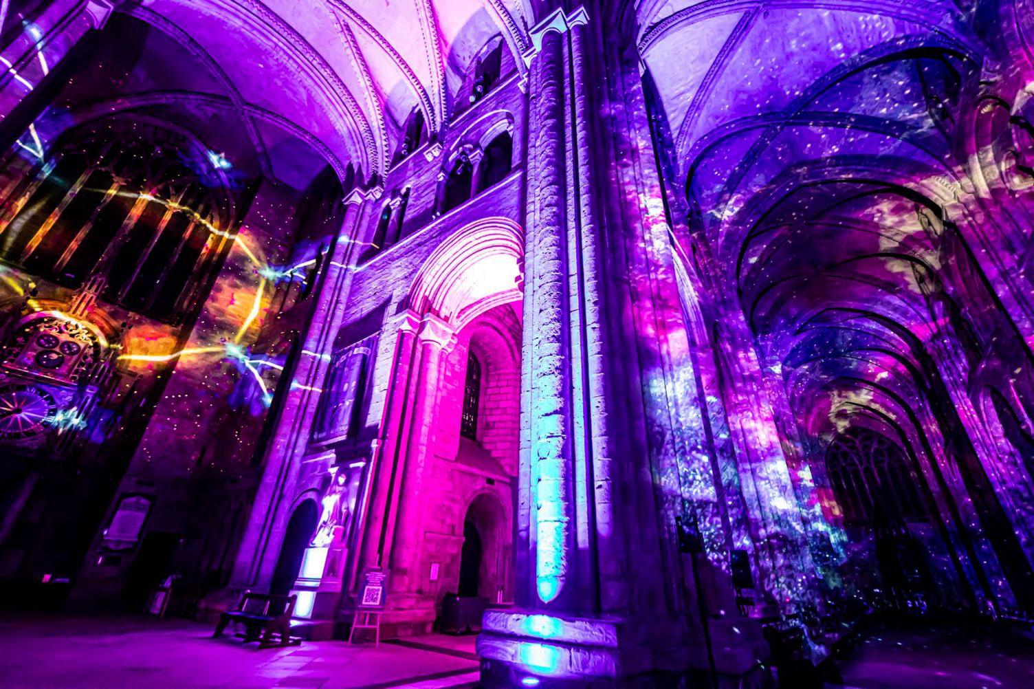 A galaxy illumination inside the cathedral. There is a bright yellow circle on the left against a backdrop of pink and purple. On the right the columns and the ceiling are lit up in a mix of blue, yellow, and pink in swirly shapes.