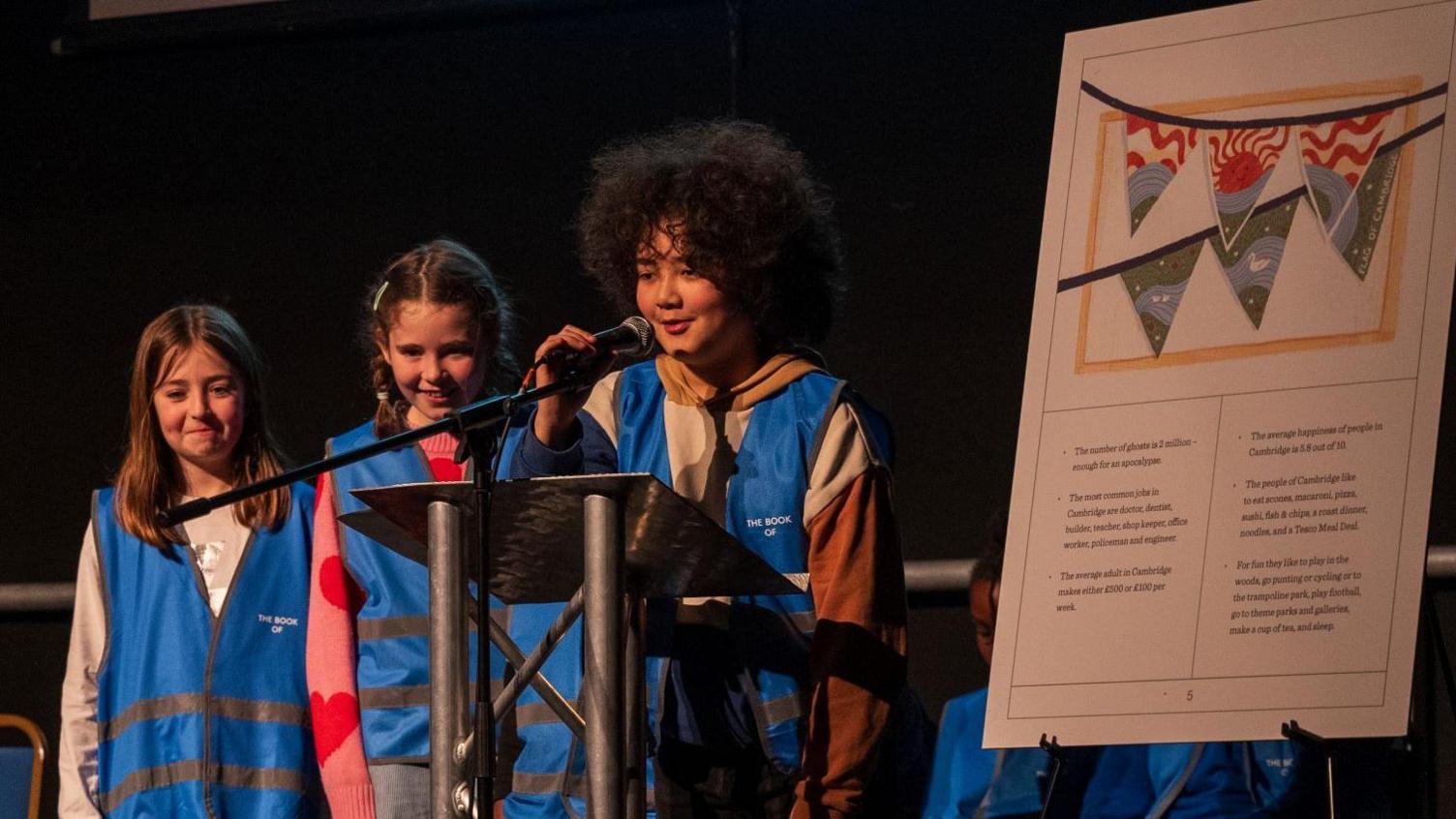 Three young girls wearing blue hi-vis vests standing on stage, next to a large version of their book. One of the girls is standing behind a lectern and speaking into a microphone. The other two look excited but have nervous smiles.