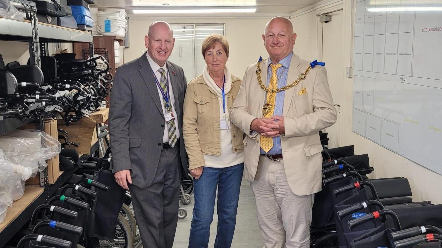 Two bald men in suits standing either side of a woman in a white hoodie and beige denim jacket. There are wheelchairs parked either side of the group.