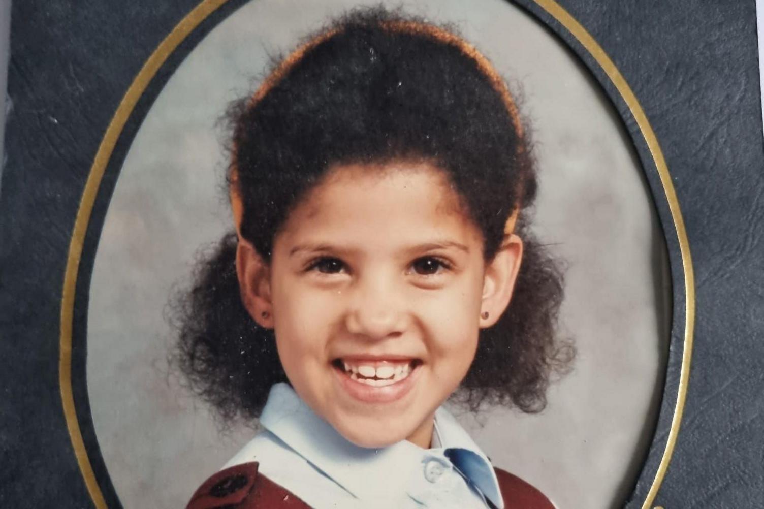 Anastasia is in her primary school uniform, posing for the professional pictures taken in school. Her curly, black Afro hair is cut to chin length and a hairband is on top of her hair.