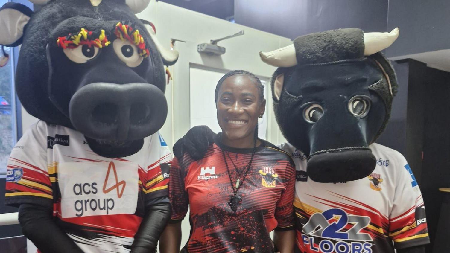 Rachel Iliya wears a red and black Bradford Bulls shirt as she is flanked by two Bull mascots from the club