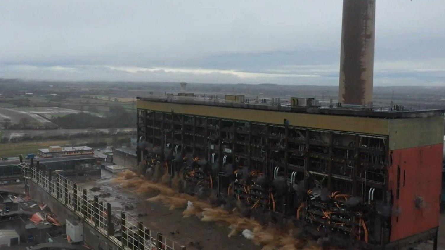 An image of a building being demolished at a decommissioned power station near Retford in Nottinghamshire. The image shows a large building with dust and rubble coming from the bottom few floors in a controlled explosion. 