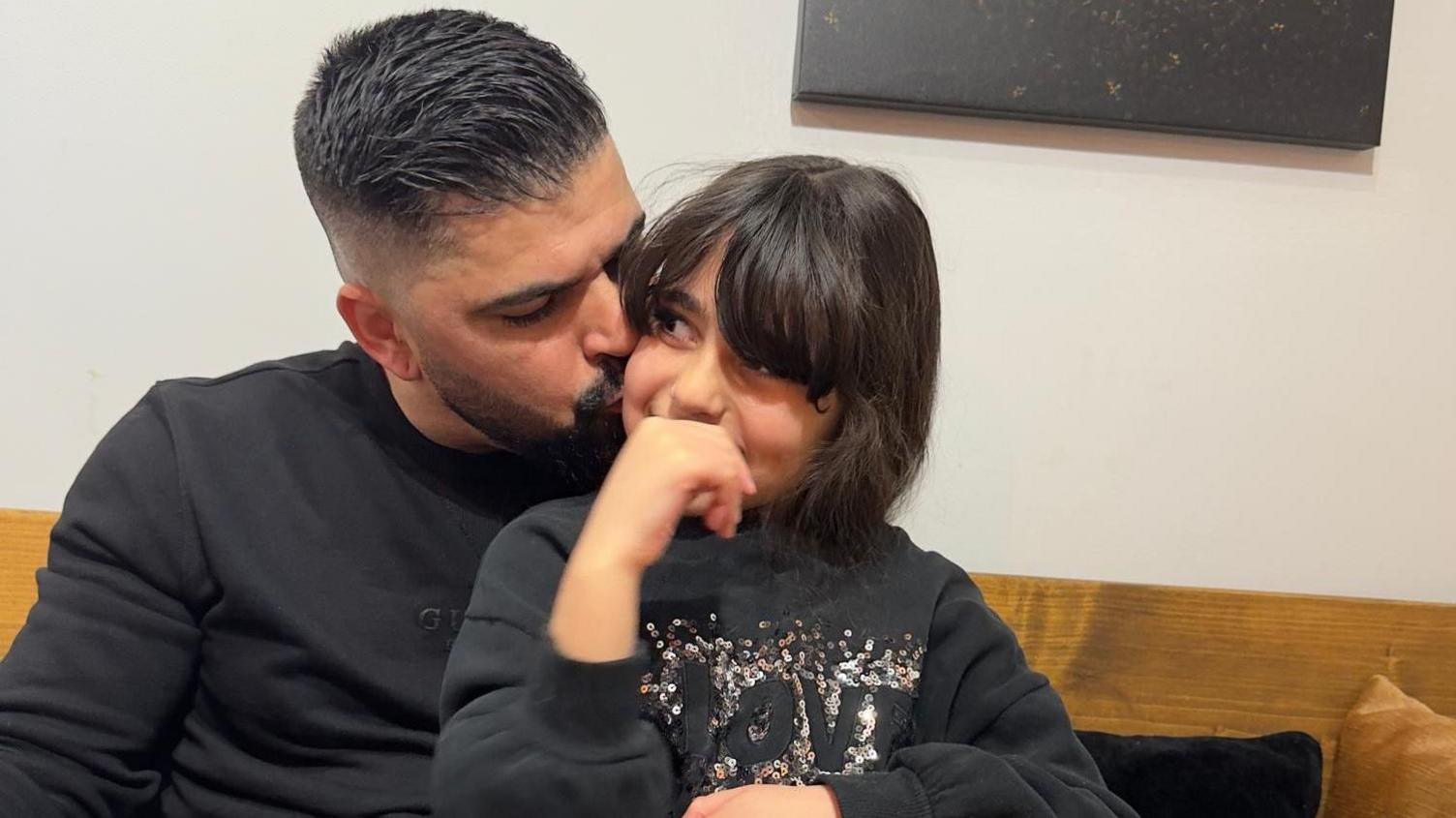 Sergio sitting with his daughter Alice in a coffee shop. Alice has dark brown hair and a fringe and is sitting on her dad's knee as he kisses her on the cheek. Both have dark sweaters on, Alice's is covered in sequins.
