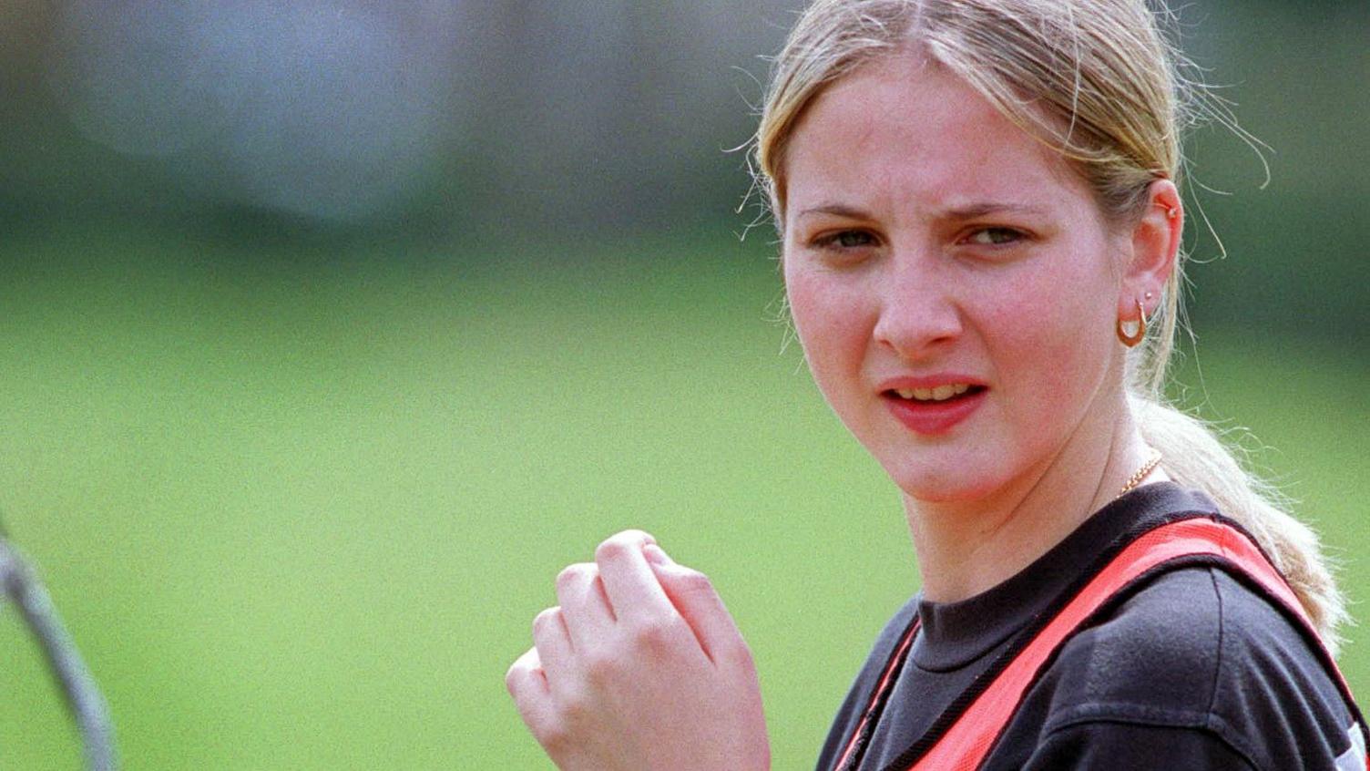 A headshot of a blonde woman looking to the right of the camera. She has straight hair tied back in a low ponytail and gold earrings and necklace. The grass of a football pitch can be seen behind her.